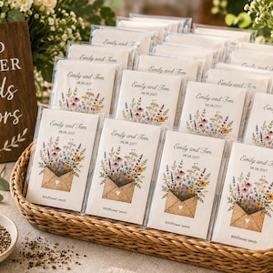 May include: A collection of rectangular seed packets in a woven basket, each featuring a printed illustration of a brown envelope overflowing with colorful wildflowers. The text reads Emily and Tom 08.08.2027 and wildflower seeds.