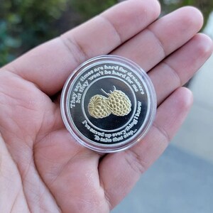 May include: A silver coin with a clear plastic case. The coin features two gold-colored raspberries and text that reads, "They say times are hard for dreams but they won't be hard for me. I've saved up everything I know to take that step..."