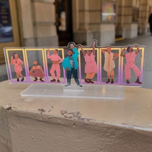 May include: A clear acrylic stand with eight colourful figures of people in various poses. The figures are printed on the acrylic and are set against a gradient background of pink and yellow.