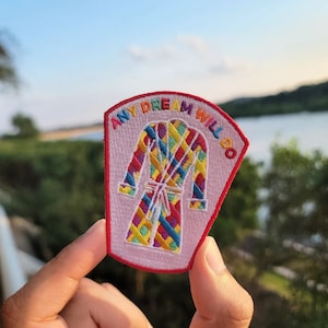 Puede incluir: Un parche bordado rosa con un diseño de bata colorido y el texto "Any Dream Will Do".