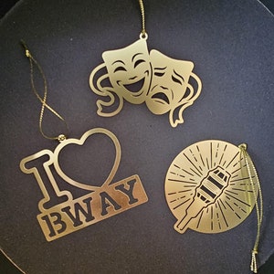 May include: Three gold-colored metal ornaments with strings. The first ornament is a pair of theater masks, one smiling and one frowning. The second ornament is a heart with the text "I LOVE BWAY". The third ornament is a circle with a microphone inside.