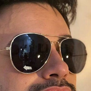 Puede incluir: Un hombre con gafas de sol aviador doradas con lentes negros. Las gafas tienen un marco de metal dorado.