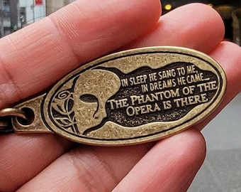 May include: A gold-colored metal keychain with a round, oval-shaped charm. The charm features a profile of a person wearing a mask and the text "IN SLEEP HE SANG TO ME, IN DREAMS HE CAME... THE PHANTOM OF THE OPERA IS THERE".