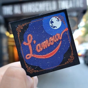 May include: A square patch with a blue background featuring a crescent moon and the text "L'amour" in red script. The patch has a black border with decorative swirls and is held in front of a theater marquee.