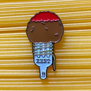 May include: A brown and red enamel pin depicting a meatball with red sauce on top, wrapped in yellow spaghetti noodles. The spaghetti is tied with a white ribbon that says "RENT" in black letters. There are two small figures at the bottom of the ribbon.
