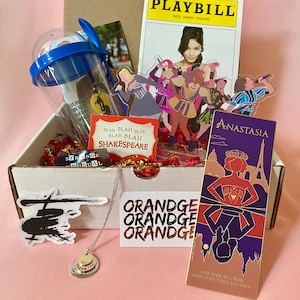 May include: A white box filled with various items, including a playbill for a Neil Simon Theatre production, a necklace with a charm that says "Not throwing away my shot", a sticker of a helicopter, a card with the words "Blah blah blah blah blah Shakespeare", and a bookmark with the words "Anastasia" and "One step at a time, one hope then another".