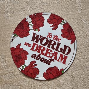 以下が含まれることがあります： 白い背景に赤い花のイラストが描かれた丸いコースター。「To the WORLD we DREAM about」というテキストが赤とマルーン色で書かれています。コースターはベージュ色の表面に置かれています。