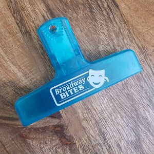 May include: A blue plastic binder clip with the text "Broadway Bites" and a white smiling face icon.