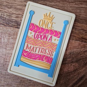 May include: A light blue and yellow bed frame with a pink and yellow patterned mattress. The text "ONCE UPON A MATTRESS" is written on the mattress with a gold crown above the word "ONCE".