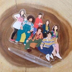 May include: A group of seven illustrated figures on a clear acrylic stand. The figures are all wearing different colorful outfits and are posed in a variety of positions. The stand is shaped like a circle and has a smooth, glossy finish.