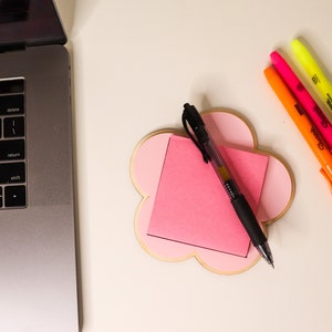 Puede incluir: Un bloc de notas rosa en forma de flor con una nota adhesiva y un bolígrafo negro en la parte superior. El bloc de notas está sobre una superficie blanca con tres resaltadores en rosa, naranja y amarillo.