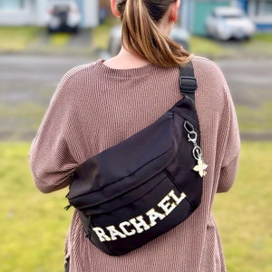 May include: A black fanny pack with a white zipper and a gold chain with a butterfly charm. The fanny pack has the name "RACHAEL" embroidered on it in white letters with gold outlining.