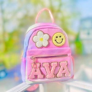 May include: A pink tie-dye mini backpack with a white flower, yellow smiley face, and the name "AVA" in pink fluffy letters.