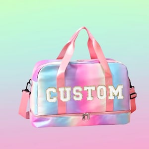 May include: A colorful duffel bag with a tie-dye pattern in shades of pink, blue, and purple. The word "CUSTOM" is embroidered in white with gold trim on the front. The bag has pink handles and a shoulder strap.