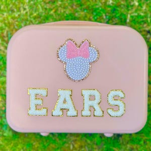 May include: A pink hard-sided case with a white and pink Minnie Mouse head with a bow and the word "EARS" in white letters with gold trim.