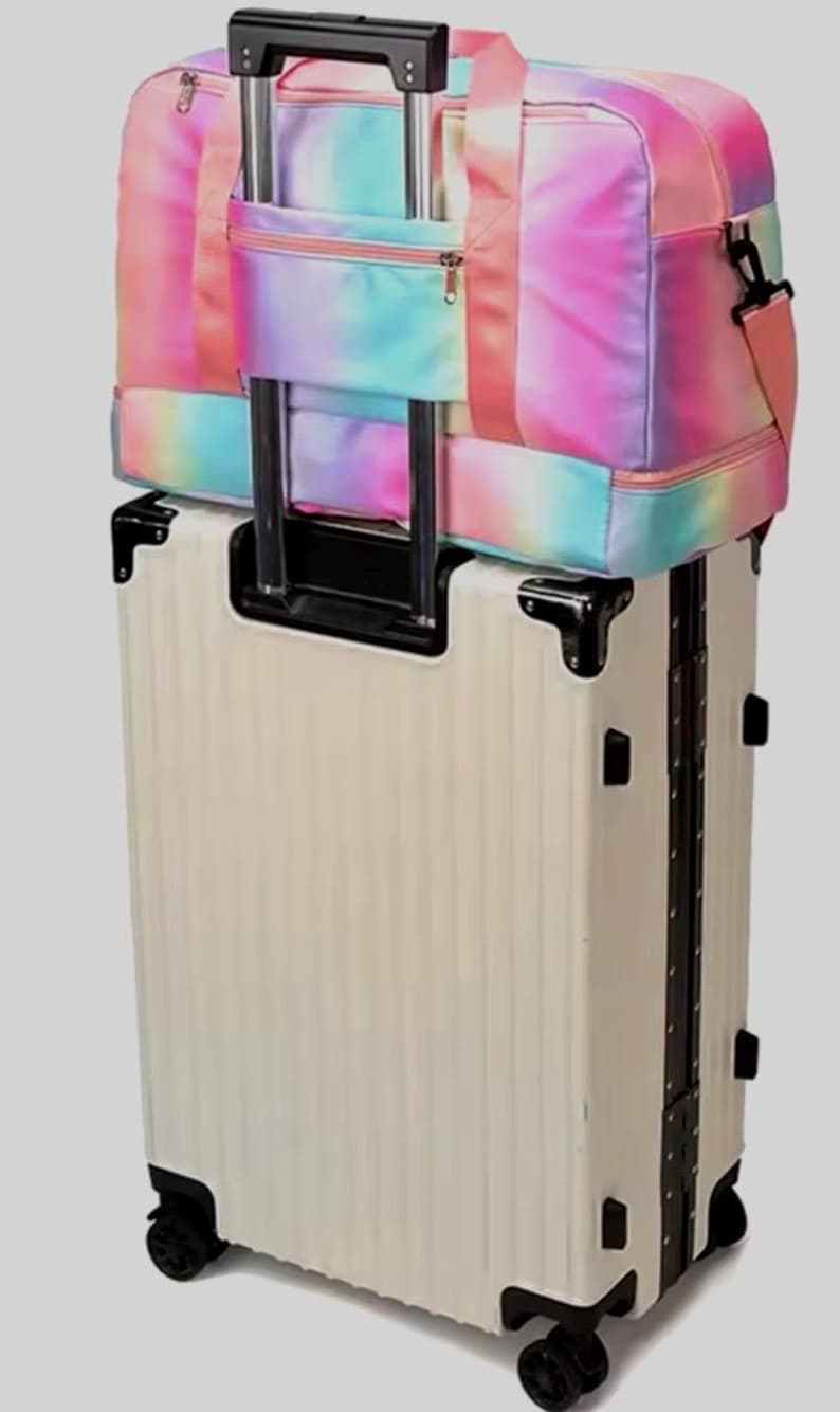 May include: A cream-colored rolling suitcase with a matching tie-dye duffel bag on top. The duffel bag features a rainbow of colors, including pink, blue, and yellow. The suitcase has black accents and wheels, perfect for travel.