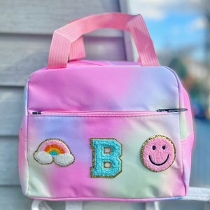 May include: A colorful lunch bag with a tie-dye effect in pink, purple, and white. The bag features a front zipper pocket and a pink handle. Patches include a rainbow, the letter "B", and a smiley face.