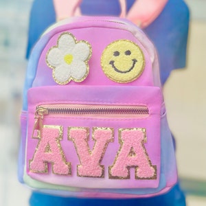 May include: A pink tie-dye backpack with a white flower, yellow smiley face, and gold glitter letters spelling "AVA".