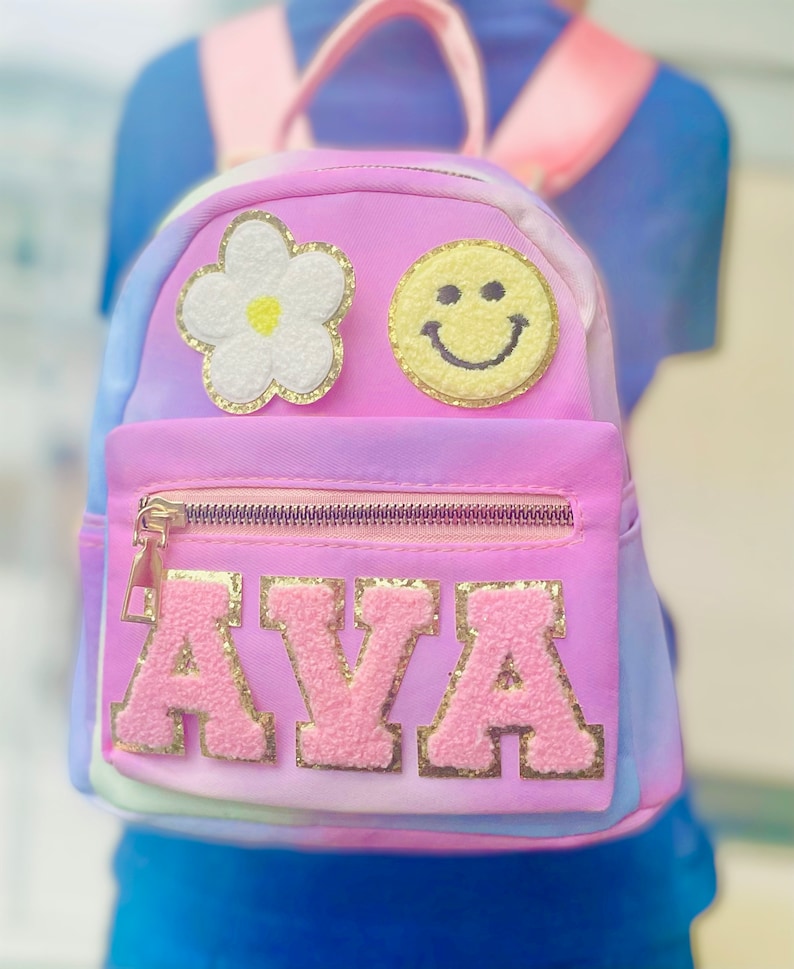 May include: A pink tie-dye backpack with a white flower, yellow smiley face, and gold glitter letters spelling "AVA".