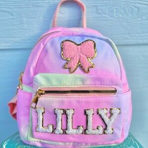 May include: A pink and blue tie-dye mini backpack with a pink bow and gold glitter accents. The backpack has a zippered pocket and the name "LILLY" is embroidered on the front in white with gold outlining.