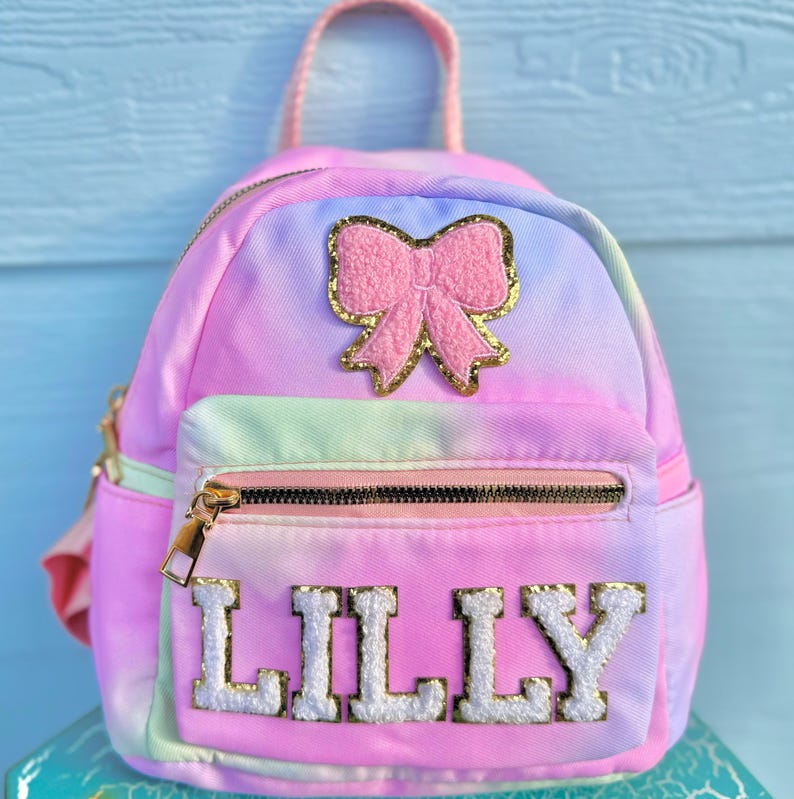 May include: A pink and blue tie-dye mini backpack with a pink bow and gold glitter accents. The backpack has a zippered pocket and the name "LILLY" is embroidered on the front in white with gold outlining.