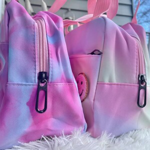 Puede incluir: Dos bolsas con estampado tie-dye en tonos rosa, morado y azul. Cada bolsa tiene un compartimento principal con cremallera y tirador negro, un peque&ntilde;o bolsillo frontal y un asa rosa. Una bolsa tiene un parche de carita sonriente.