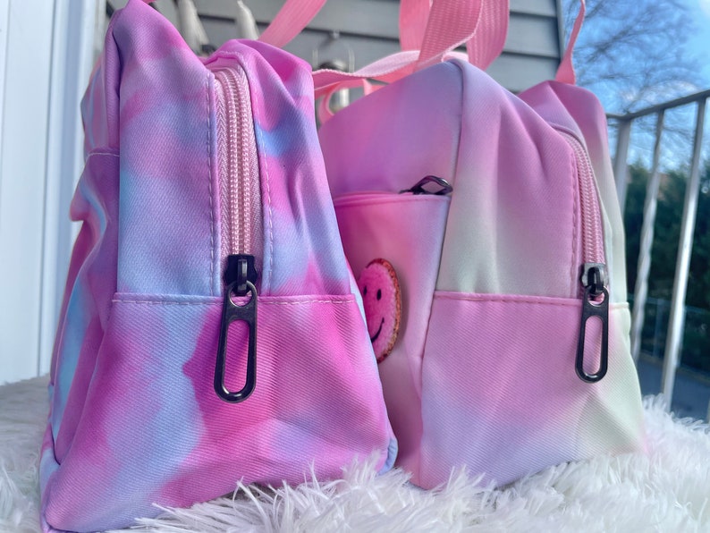 Puede incluir: Dos bolsas con estampado tie-dye en tonos rosa, morado y azul. Cada bolsa tiene un compartimento principal con cremallera y tirador negro, un peque&ntilde;o bolsillo frontal y un asa rosa. Una bolsa tiene un parche de carita sonriente.