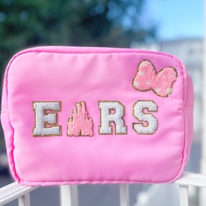 May include: Pink zippered pouch with the word "EARS" embroidered in white with a pink castle in the middle of the word. A pink bow with white polka dots is on the top right corner of the pouch.