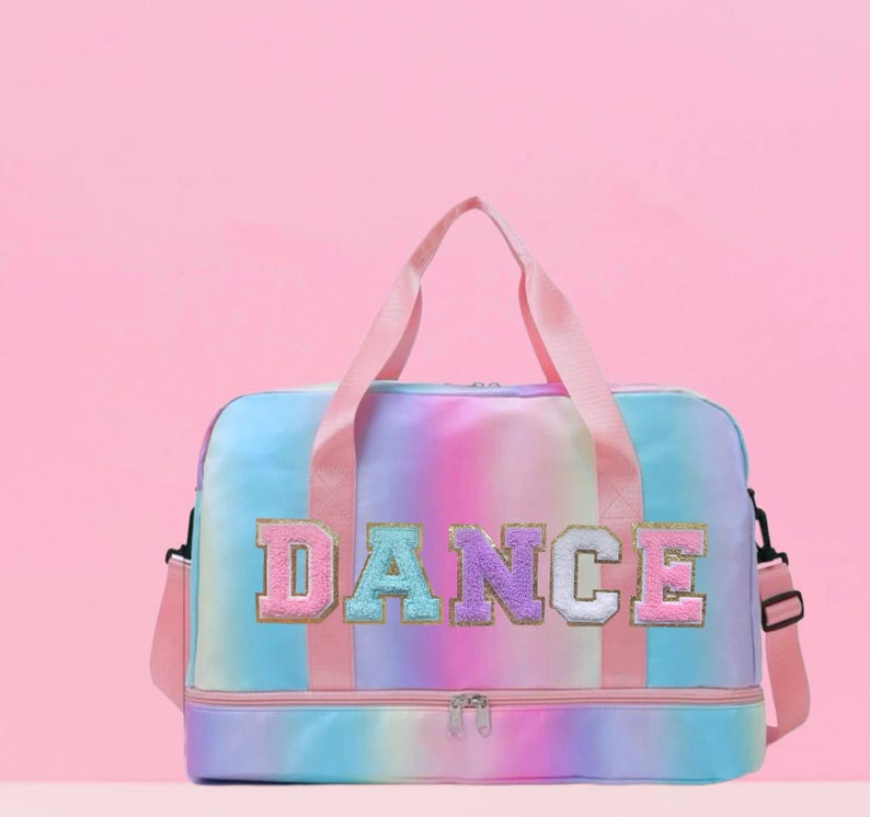May include: A colorful dance bag with a rainbow tie-dye pattern. The word "DANCE" is embroidered across the front in pink, blue, purple, and gold letters. The bag has pink handles and a detachable shoulder strap.