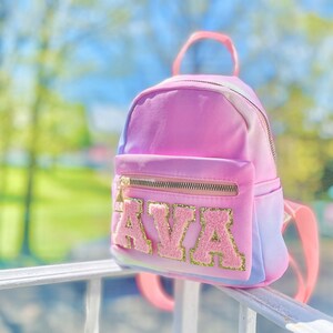 May include: A pink and blue ombre mini backpack with a gold zipper and the name "AVA" in pink and gold glitter letters.