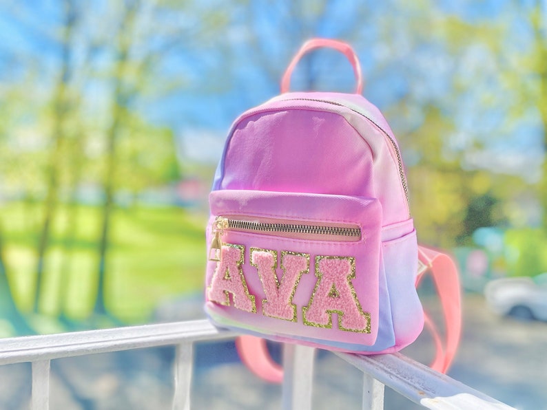 May include: A pink and blue ombre mini backpack with a gold zipper and the name "AVA" in pink and gold glitter letters.