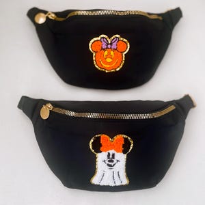 May include: Two black fanny packs with gold zippers and pulls. One has an orange Minnie Mouse face with a purple bow, the other has a white ghost Minnie Mouse face with an orange bow. The fanny packs are on a white background.