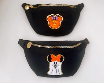 May include: Two black fanny packs with gold zippers and pulls. One has an orange Minnie Mouse face with a purple bow, the other has a white ghost Minnie Mouse face with an orange bow. The fanny packs are on a white background.
