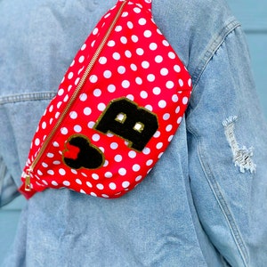 May include: Red fanny pack with white polka dots and a gold zipper. The bag features a black patch with a gold-trimmed letter "B" and a black heart. The strap is red.