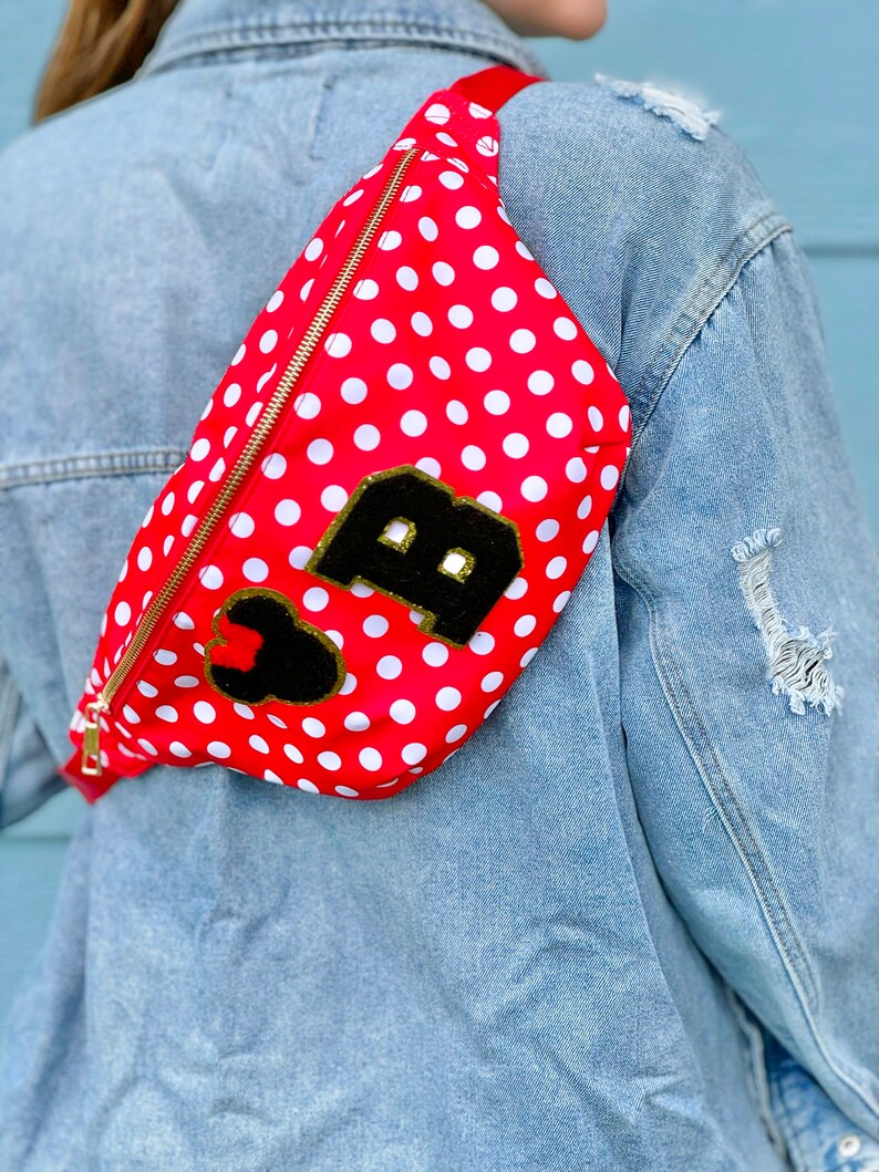 May include: Red fanny pack with white polka dots and a gold zipper. The bag features a black patch with a gold-trimmed letter "B" and a black heart. The strap is red.