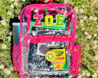 May include: A clear pink backpack with the name "ZOE" in green and yellow letters. The backpack contains a notebook, crayons, and a smiley face patch. A water bottle is visible in the side pocket.