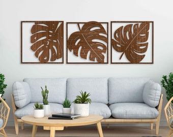 May include: Three walnut wood wall decor pieces featuring a cutout design of monstera leaves. The pieces are arranged in a row on a white wall above a light gray sofa.
