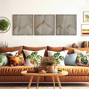 May include: A brown leather sofa with multiple throw pillows in various patterns and colors, including green, orange, and blue. The sofa is in front of a white wall with three wooden wall decor pieces featuring a geometric design.