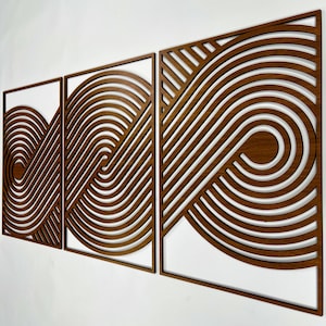 Op de afbeelding: Set van drie houten wandpanelen. Elk paneel heeft een uniek ontwerp van gebogen, parallelle lijnen in een warme bruine tint, waardoor een abstract, geometrisch patroon ontstaat tegen een witte achtergrond. De panelen zijn ingelijst.