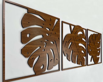 May include: Three wooden wall decor pieces featuring a cutout design of tropical leaves. The leaves are a dark brown color and are arranged in a staggered pattern.