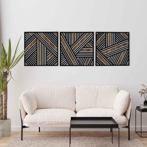 May include: Three black framed wall art pieces with a geometric pattern of brown and white stripes. The art is displayed on a white wall above a white couch and a black coffee table.