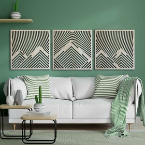 May include: Three wooden wall art panels with a geometric mountain design. The panels are in a light wood tone and feature a pattern of overlapping lines and shapes. The panels are arranged in a row on a green wall above a white sofa.
