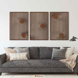 May include: Three framed wooden wall art pieces with a wavy, striped design and circular cutouts. The frames are a dark brown, and the artwork is mounted above a gray sofa with decorative pillows.