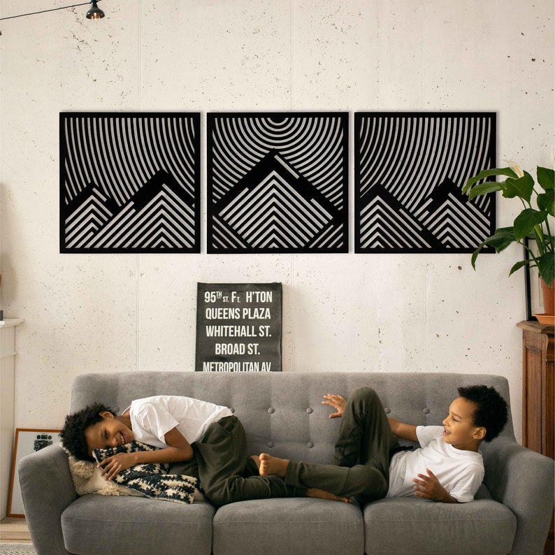 May include: Three black and white geometric wall art pieces with a mountain design. The art is hung on a white wall above a gray couch with two children sitting on it.