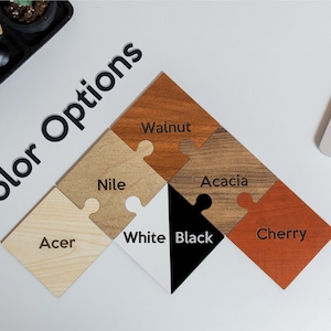 May include: A collection of wooden puzzle pieces in various colors, including walnut, acacia, cherry, and black. The text "Color Options" is displayed in black, along with the names of each color on the puzzle pieces.