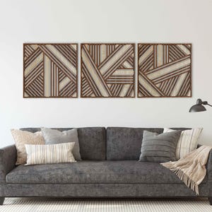 May include: Three brown wood wall decor panels with a geometric pattern of intersecting lines. The panels are arranged horizontally and are hanging on a white wall above a gray couch with pillows.