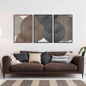 May include: Three framed wall art pieces with concentric circle designs in shades of brown, black, and white. The artwork hangs above a brown sofa with cushions and a striped rug. A floor lamp and potted plant are also visible.