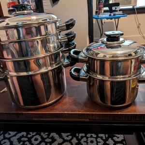 May include: A set of stainless steel cookware featuring a large stack of three pots with black handles and a matching lid, alongside a smaller two-piece pot set with a lid. The polished metal surfaces reflect the surrounding environment clearly.