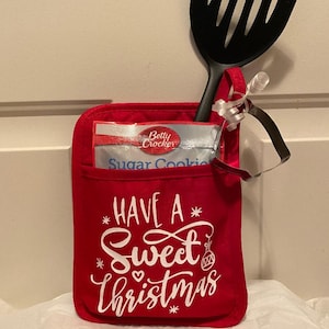 May include: Red Christmas-themed oven mitt with the text "Have a Sweet Christmas" in white. The mitt has a pocket with a packet of Betty Crocker Sugar Cookies inside. A black spatula and a heart-shaped cookie cutter are sticking out of the pocket.