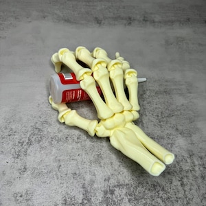 May include: A cream-coloured, plastic skeleton hand holding a red and white bottle. The hand is resting on a grey surface. The bottle has a white cap and red label.